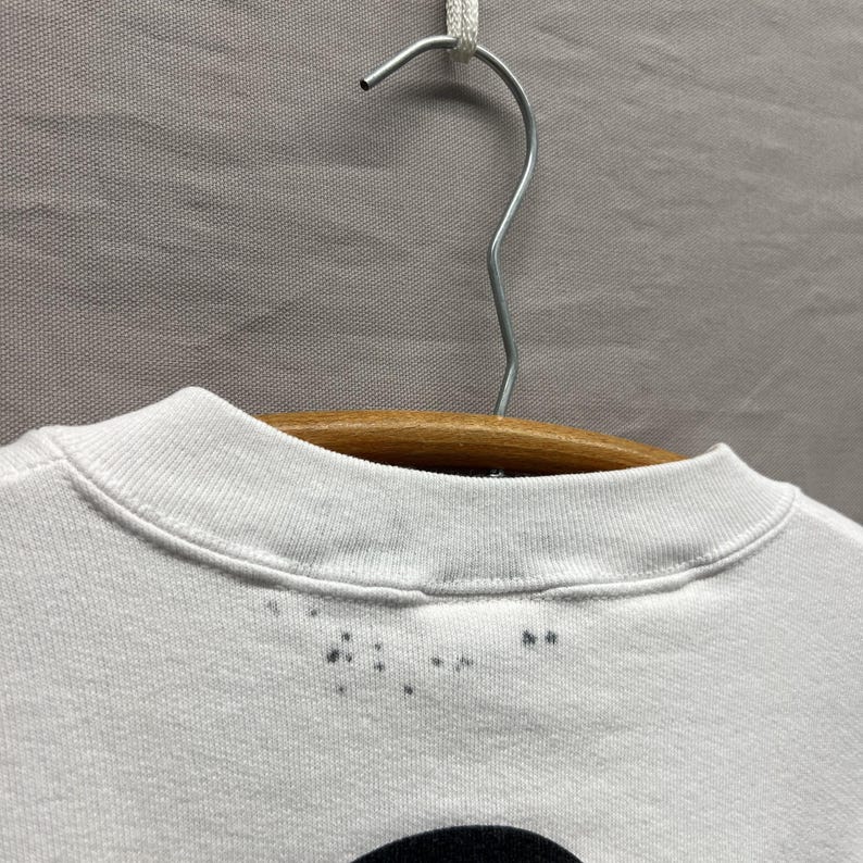 May include: A white crew neck t-shirt hanging on a wooden hanger. The collar features a ribbed neckline with a series of small, dark grey spots or stains scattered along the front edge just below the seam of the collar, visible against the fabric.