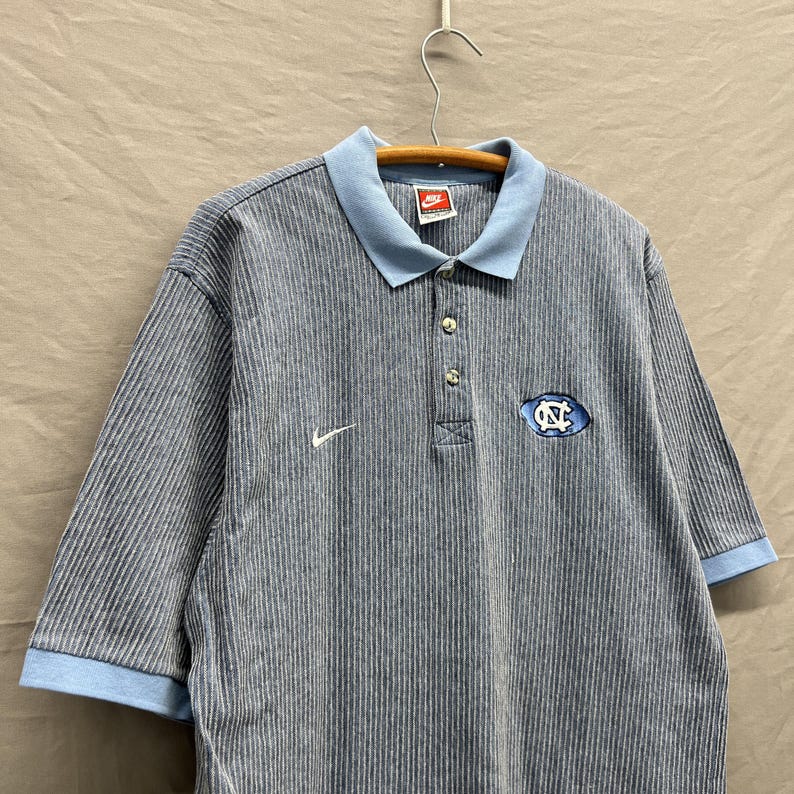 Puede incluir: Un polo de manga corta con un patr&oacute;n de rayas finas grises y blancas, con cuello y pu&ntilde;os de color azul claro. La prenda presenta un logotipo Nike blanco en el pecho derecho y un logotipo azul y blanco de Carolina del Norte en el izquierdo.