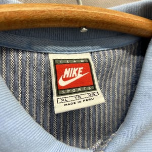 Puede incluir: Una camisa azul claro con un patr&oacute;n de rayas verticales texturizadas. El cuello interior presenta una etiqueta blanca con el logotipo rojo y negro de Nike Team Sports, la talla XL y el texto Made in Peru. La prenda est&aacute; en una percha de madera.