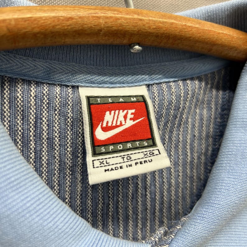 Puede incluir: Una camisa azul claro con un patr&oacute;n de rayas verticales texturizadas. El cuello interior presenta una etiqueta blanca con el logotipo rojo y negro de Nike Team Sports, la talla XL y el texto Made in Peru. La prenda est&aacute; en una percha de madera.