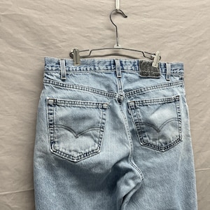 May include: Light blue denim jeans hanging on a metal hanger. The jeans have two back pockets with a curved design and a leather patch. The denim is faded, showing signs of wear.