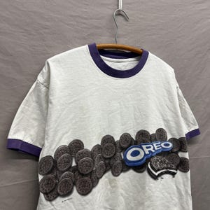 May include: A white short-sleeve t-shirt with a dark purple crew neckline and sleeve cuffs. The front features a graphic print of numerous dark brown Oreo cookies and the blue and white Oreo logo, with one cookie shown split in half.
