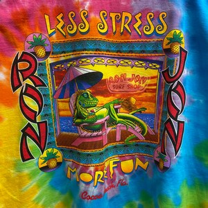 May include: A tie-dye t-shirt with a cartoon iguana lounging on a chair under a beach umbrella. The shirt features the text "Less Stress More Fun" and "Ron Jon Surf Shop".