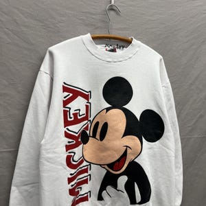 May include: A white long-sleeve crewneck sweatshirt featuring a large graphic of Mickey Mouse on the front. The word MICKEY is printed vertically in bold red letters next to the character. The interior collar tag has the handwritten text Mickey.