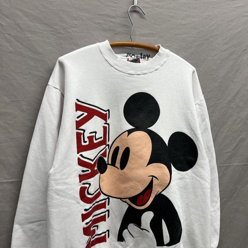 May include: A white long-sleeve crewneck sweatshirt featuring a large graphic of Mickey Mouse on the front. The word MICKEY is printed vertically in bold red letters next to the character. The interior collar tag has the handwritten text Mickey.