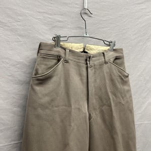 May include: A pair of light brown trousers featuring a high-waisted design with belt loops, a front zipper closure, and two deep side pockets. The interior waistband is lined with a cream-colored fabric, and the garment hangs on a metal hanger.
