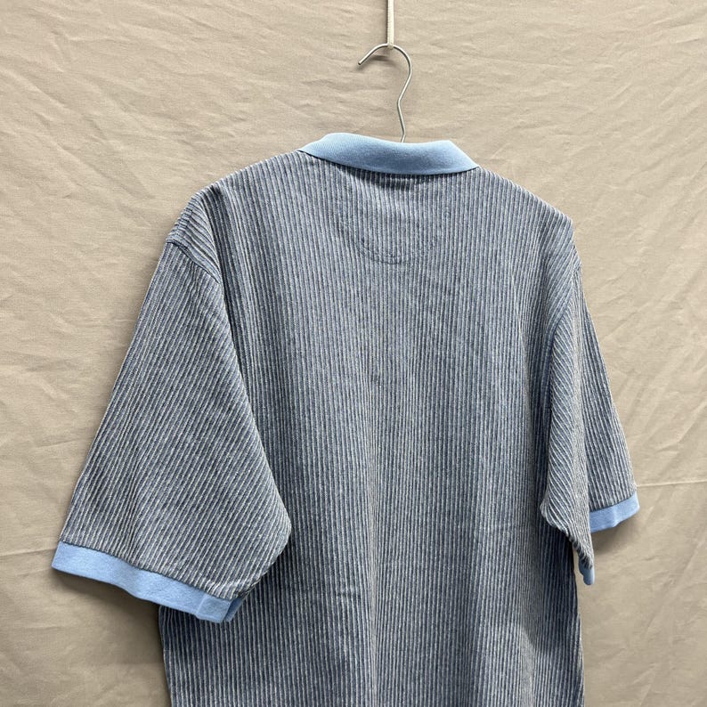 Puede incluir: Un polo de manga corta con un patr&oacute;n de rayas finas en gris oscuro y blanco. La prenda incluye un cuello de color azul claro s&oacute;lido y pu&ntilde;os a juego en el mismo tono. La camisa est&aacute; colgada de un gancho met&aacute;lico sobre un fondo neutro.