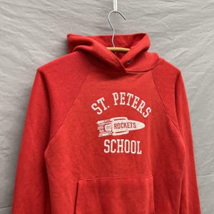 May include: A red hooded sweatshirt with the text "ST. PETERS ROCKETS SCHOOL" in white. The graphic includes a rocket ship. The sweatshirt has a front pocket and is hanging on a hanger.
