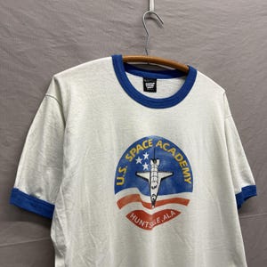 May include: A white ringer t-shirt with blue trim on the collar and sleeves. The front features a circular graphic with a space shuttle, three white stars, and the text U.S. SPACE ACADEMY HUNTSVILLE, ALA in yellow, blue, and red ink.