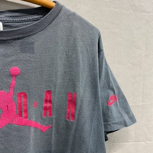 May include: A vintage grey t-shirt with a pink Jordan logo and the word "JORDAN" in pink lettering. The Nike logo is on the right sleeve.