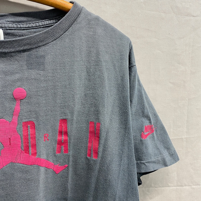 May include: A vintage grey t-shirt with a pink Jordan logo and the word "JORDAN" in pink lettering. The Nike logo is on the right sleeve.