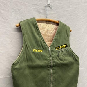 May include: Olive green military vest with a full-length zipper and a V-neck. The vest has a quilted, tan lining and a small pocket on the right side. The text "CALSON" is embroidered in yellow on the left side, and "U.S. ARMY" is on the right.