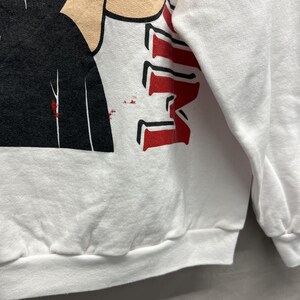 May include: A white long-sleeve sweatshirt featuring a graphic print with a black shape, a tan-colored section, and bold red lettering. The design includes small red speckles near the black area, set against the plain white fabric of the garment.