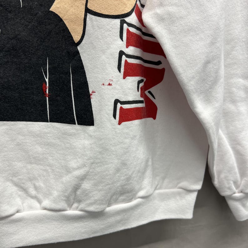 May include: A white long-sleeve sweatshirt featuring a graphic print with a black shape, a tan-colored section, and bold red lettering. The design includes small red speckles near the black area, set against the plain white fabric of the garment.