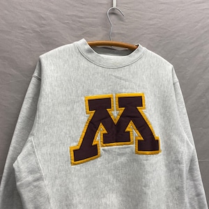 May include: A heather gray sweatshirt with a large, appliqued "M" logo. The logo features a maroon letter outlined in gold. The sweatshirt has a crew neck and long sleeves, and is hanging on a wooden hanger.