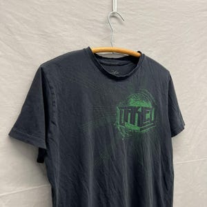 May include: Black short-sleeve t-shirt with the word "OAKLEY" in green block letters inside a green circle. The shirt is hanging on a wooden hanger against a light gray background.