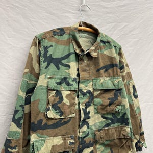 May include: Camouflage pattern jacket with multiple pockets. The jacket features a mix of green, brown, and black hues. It is hanging on a wooden hanger against a neutral background. The jacket has a collared neckline and button-down front.