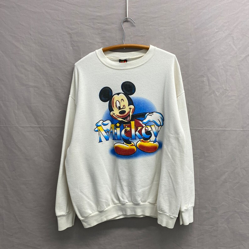 May include: White sweatshirt with a colorful graphic of Mickey Mouse winking and the word "Mickey" printed in blue, yellow, and red.