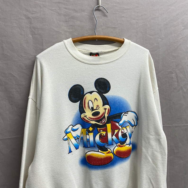 May include: White sweatshirt with a colorful graphic of Mickey Mouse winking and the word "Mickey" printed in blue, yellow, and red.