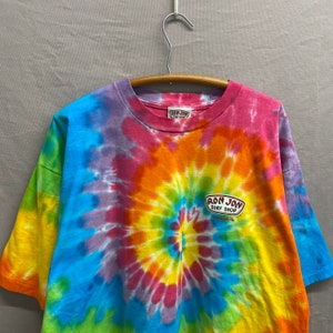 May include: A tie-dye t-shirt with a rainbow swirl pattern. The shirt features a vintage Ron Jon Surf Shop logo in the center.