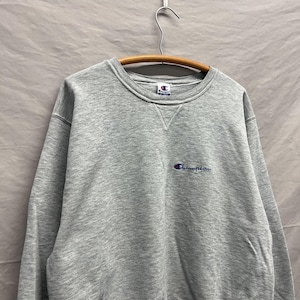 May include: A heather gray Champion sweatshirt with a crew neck and long sleeves. The Champion logo is embroidered in blue on the chest. The sweatshirt is hanging on a wooden hanger.