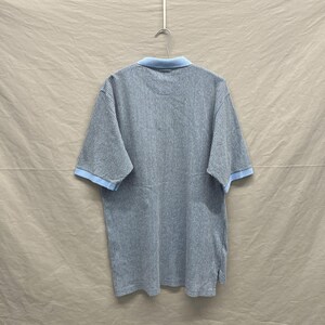 Puede incluir: Un polo de manga corta con un patr&oacute;n de rayas verticales finas en azul oscuro y blanco. La prenda incluye un cuello de color azul claro s&oacute;lido y ribetes a juego en el mismo tono azul claro en los pu&ntilde;os, colgado en una percha de metal.