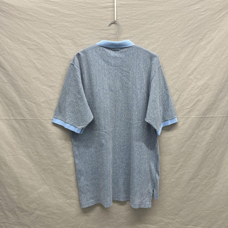 Puede incluir: Un polo de manga corta con un patr&oacute;n de rayas verticales finas en azul oscuro y blanco. La prenda incluye un cuello de color azul claro s&oacute;lido y ribetes a juego en el mismo tono azul claro en los pu&ntilde;os, colgado en una percha de metal.