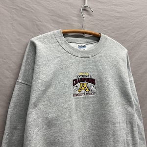May include: A heather grey crewneck sweatshirt featuring an embroidered logo on the chest that reads NCAA NATIONAL CHAMPIONS 2002 MINNESOTA HOCKEY with a gold letter M and crossed hockey sticks. The garment has a ribbed collar and cuffs.