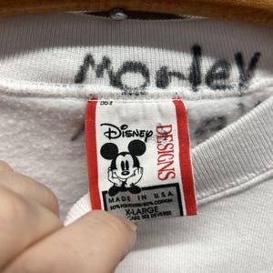 May include: A white sweatshirt featuring a Disney Designs brand tag with a Mickey Mouse graphic. The tag includes the text Disney Designs, Made in U.S.A., 50% Polyester/50% Cotton, and X-Large. The word Morley is handwritten in black ink on the collar.