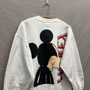 May include: A white long-sleeve sweatshirt featuring a large graphic of Mickey Mouse on the back. The character is depicted in black and tan, positioned next to the vertical red text that reads MICKEY. The garment is displayed on a hanger.