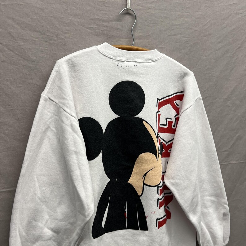 May include: A white long-sleeve sweatshirt featuring a large graphic of Mickey Mouse on the back. The character is depicted in black and tan, positioned next to the vertical red text that reads MICKEY. The garment is displayed on a hanger.