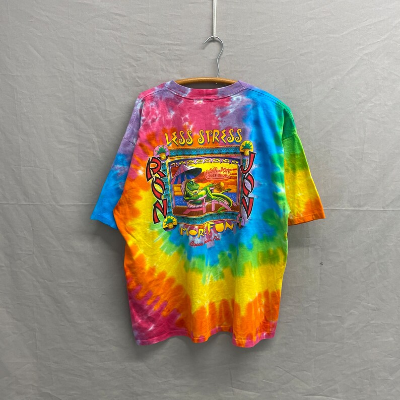 May include: A tie-dye t-shirt with a cartoon illustration of a lizard relaxing on a beach chair. The shirt has the text "Less Stress More Fun" and "Ron Jon Surf Shop" printed on it.