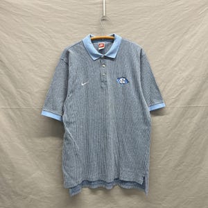 Puede incluir: Un polo de manga corta con un patr&oacute;n de rayas finas en gris y blanco, con cuello y pu&ntilde;os en color azul claro. La prenda presenta un logotipo Nike blanco en el pecho derecho y un parche con el logotipo NC en azul y blanco a la izquierda.