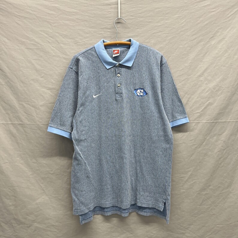 Puede incluir: Un polo de manga corta con un patr&oacute;n de rayas finas en gris y blanco, con cuello y pu&ntilde;os en color azul claro. La prenda presenta un logotipo Nike blanco en el pecho derecho y un parche con el logotipo NC en azul y blanco a la izquierda.
