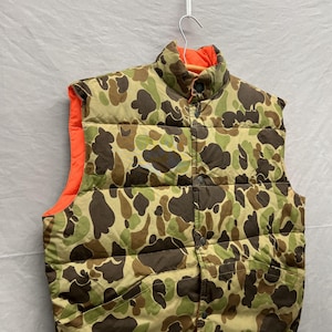May include: A camouflage-patterned vest with a high collar and button closure. The vest features a brown, green, and tan pattern on the exterior and a bright orange lining. It is hanging on a white hanger.