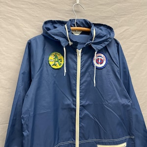 May include: A vintage navy blue windbreaker jacket with a hood and a white zipper. The jacket features patches for the Oakland A's and Minnesota Twins baseball teams. The jacket is hanging on a metal hanger.
