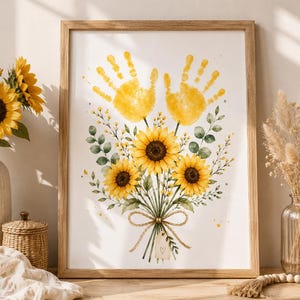 Könnte beinhalten: Gerahmter Aquarelldruck mit einem Sonnenblumenstrauß und zwei gelben Handabdrücken. Der Strauß ist mit Bindfaden gebunden und enthält grüne Blätter und kleine gelbe Blüten. Der Rahmen besteht aus hellbraunem Holz.