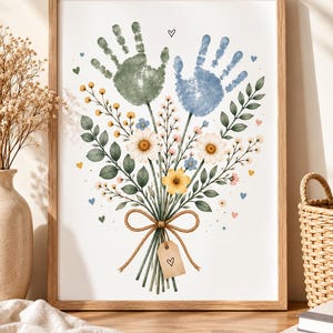 Puede incluir: Una lámina artística enmarcada que muestra un ramo de flores con dos huellas de manos en la parte superior, una en verde salvia y otra en azul empolvado. Los tallos están atados con un lazo de cuerda marrón y una etiqueta con un corazón.