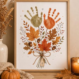 Op de afbeelding: Een ingelijste kunstdruk met een boeket van herfstbladeren, eikels en bessen, bekroond met twee handafdrukken in olijfgroen en oranje. De stelen zijn samengebonden met een touwtje en een klein rechthoekig label met een hartje.