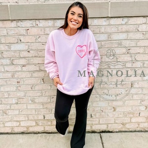 Puede incluir: Una sudadera rosa con un gr&aacute;fico de coraz&oacute;n rosa que dice "Coffee". La modelo lleva pantalones negros y est&aacute; de pie frente a una pared de ladrillo.