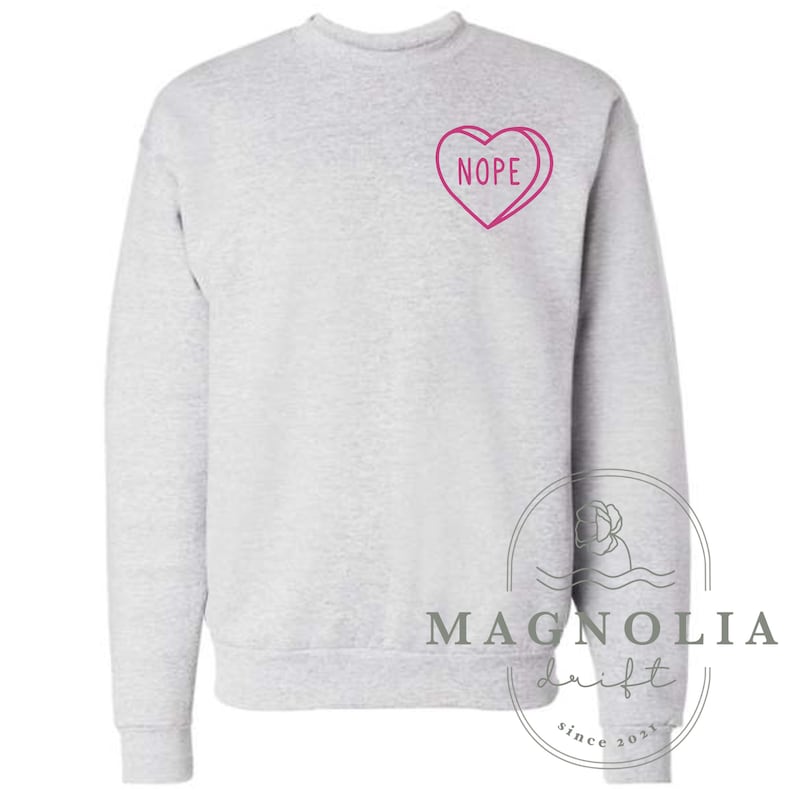 Puede incluir: Una sudadera gris con un dise&ntilde;o de caramelo en forma de coraz&oacute;n rosa que dice "NOPE".