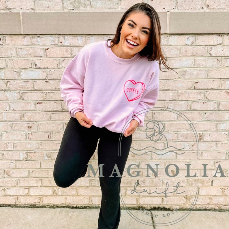 Puede incluir: Una sudadera rosa con un gr&aacute;fico de coraz&oacute;n rojo que dice "Coffee". La modelo lleva pantalones negros y sonr&iacute;e.