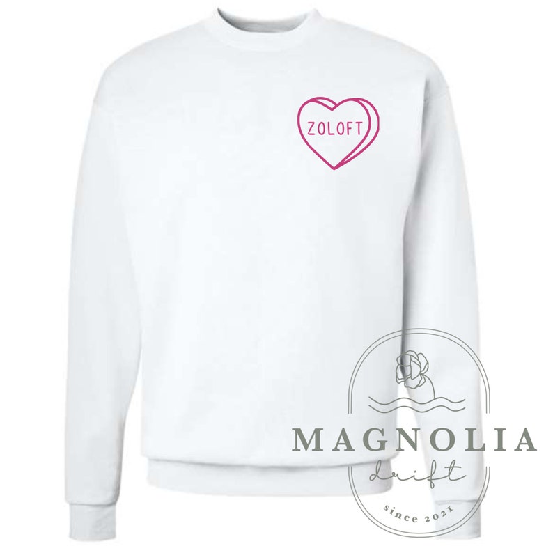 Puede incluir: Sudadera blanca con un gr&aacute;fico en forma de coraz&oacute;n rosa que dice "Zoloft". La sudadera tiene mangas largas y cuello redondo.