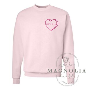Puede incluir: Sudadera rosa con un gr&aacute;fico de coraz&oacute;n rosa que dice "Chick-fil-A". La sudadera es de cuello redondo y tiene mangas largas.