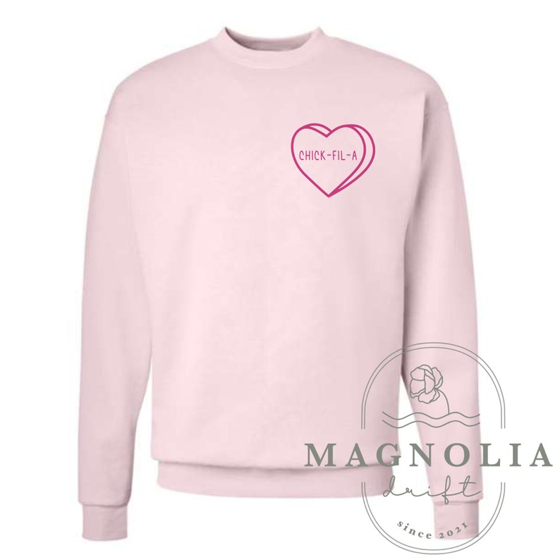 Puede incluir: Sudadera rosa con un gr&aacute;fico de coraz&oacute;n rosa que dice "Chick-fil-A". La sudadera es de cuello redondo y tiene mangas largas.