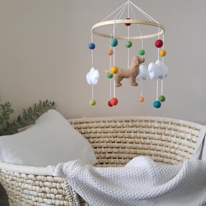 May include: A wooden baby mobile with a felt dog, clouds, and colorful balls. The mobile is hanging above a wicker basket with a white blanket.