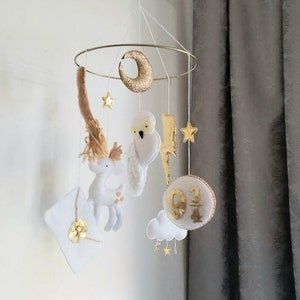 Op de afbeelding: Een witte en gouden babymobiel met een halve maan, sterren, een witte uil, een hert, een brief en een wolk met het nummer "9 3/4" erop.