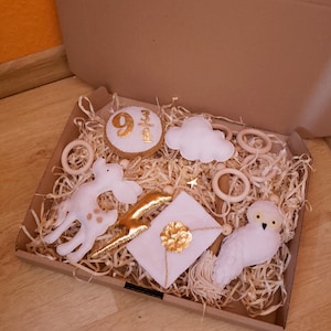Pode incluir: Uma caixa de cartão castanho cheia de brinquedos de feltro branco e dourado para bebés. Os brinquedos incluem uma nuvem, uma girafa, uma coruja, uma estrela, uma carta e um círculo com os números 9, 3 e 4.