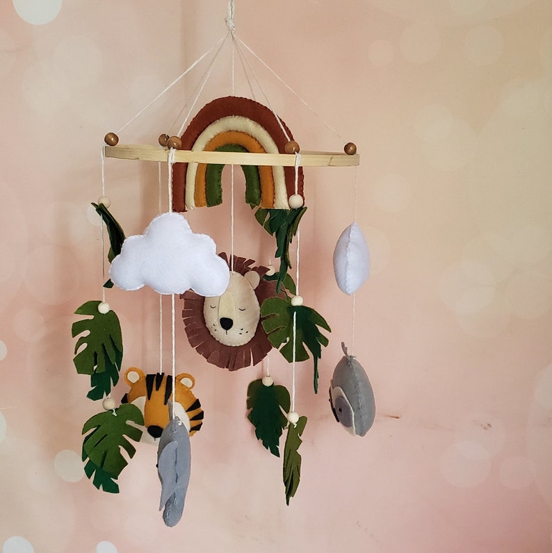 May include: A wooden baby mobile with a rainbow, cloud, lion, tiger, owl, and leaves. The mobile is made of felt and is hung from a wooden ring.