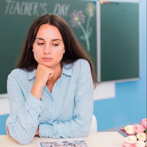 May include: A person with long dark hair wearing a light blue button-down shirt, resting their chin on their hand. Behind them is a dark green chalkboard with the text "TEACHER'S DAY" written in white chalk and a drawing of a flower.